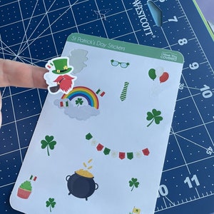 May include: A sheet of St. Patrick's Day stickers featuring a leprechaun, rainbow, shamrocks, a pot of gold, cupcakes, and other festive designs. The stickers are on a white background.