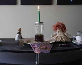 Op de afbeelding: Een mystiek stilleven met een brandende groene kaars op een pot gevuld met vloeistof en kruiden. Een pop, tarotkaarten en kopjes staan op een donkere tafel, wat een mystieke sfeer creëert.