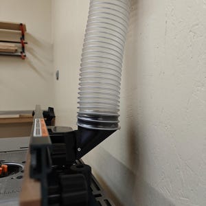 May include: A clear, flexible dust collection hose is connected to a black angled fitting, mounted on a woodworking table. The hose is ribbed and extends upwards. The table has a wooden edge and black components.