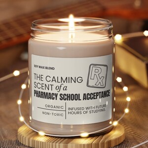 May include: A soy wax candle with a brown label that reads "The Calming Scent of a Pharmacy School Acceptance" with the text "Organic Non-Toxic" and "Infused with future hours of studying." The candle is lit and sitting on a wooden coaster.