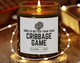 May include: A lit candle in a glass jar with a white label that reads "Smells Better Than Your Cribbage Game." The candle sits on a wooden coaster, surrounded by string lights. The candle is labeled "All Natural | 40 HRS."