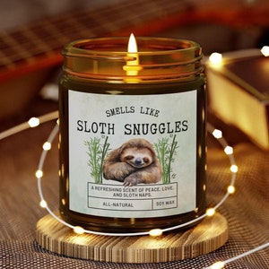 May include: A brown glass candle with a white label that reads "Smells Like Sloth Snuggles". The label features a cartoon sloth and the text "A refreshing scent of peace, love, and sloth naps." The candle is on a wooden coaster and surrounded by white fairy lights.
