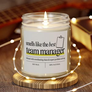 May include: A white candle with a black and white label that reads "smells like the best team manager". The label also says "Infused with coordinating chaos & expert problem solving", "Soy Wax", and "100% Natural".