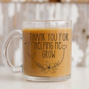 Könnte beinhalten: Eine durchsichtige Glas-Tasse mit einer braunen Flüssigkeit darin. Die Tasse hat einen Henkel und ein schwarzes Design mit dem Text "Danke, dass du mir beim Wachsen hilfst" sowie einem Stethoskop und einem Herzen.