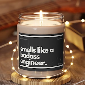 Pode incluir: Uma vela de cera de soja marrom com um rótulo preto que diz "smells like a badass engineer". A vela está acesa e está sobre um descanso de copo de madeira.
