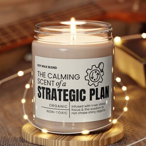 May include: A soy wax candle with a white label that reads "THE CALMING SCENT of a STRATEGIC PLAN". The label also includes the text "ORGANIC NON-TOXIC" and "Infused with laser sharp focus & the wisdom to not chase shiny objects".