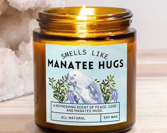 May include: A brown glass candle jar with a white label that reads "Smells Like Manatee Hugs". The label features a cartoon manatee and the text "A refreshing scent of peace, love, and manatee hugs. All-natural soy wax."