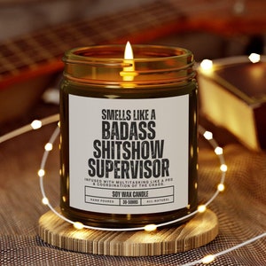 Puede incluir: Una vela de cera de soja encendida en un tarro de vidrio con la etiqueta "Smells Like a Badass Shitshow Supervisor". La vela está sobre un posavasos de madera, rodeada de luces de cadena. En el fondo, una guitarra y una caja pequeña.
