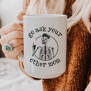May include: White ceramic mug with a black and white illustration of a skeleton holding a cup. The text on the mug reads "Go ask your other mom".