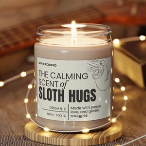 May include: A soy wax candle in a clear glass jar with a brown label that reads "The Calming Scent of Sloth Hugs". The label also says "Organic, Non-Toxic" and "Made with peace, love, and gentle snuggles". The candle is lit and sitting on a wooden coaster.