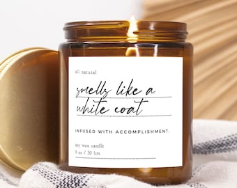Könnte beinhalten: Ein braunes Glasgefäß für eine Kerze mit einem goldenen Deckel. Das Etikett lautet "all natural smells like a white coat INFUSED WITH ACCOMPLISHMENT. soy wax candle 9 oz / 50 hrs"