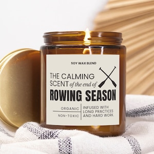 Puede incluir: Una vela de vidrio marrón con una etiqueta blanca que dice "SOY WAX BLEND THE CALMING SCENT of the end of ROWING SEASON ORGANIC NON-TOXIC INFUSED WITH LONG PRACTICES AND HARD WORK."