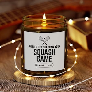 Op de afbeelding: Een brandende kaars in een glazen pot met een wit etiket met de tekst "Smells Better Than Your Squash Game". De kaars staat op een houten onderzetter, omringd door lichtslingers. De kaars is volledig natuurlijk en heeft een brandtijd van 40 uur.