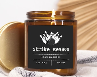 May include: A brown glass candle jar with a black label that says "strike season" and features a white illustration of bowling pins and a bowling ball. The label also says "100% natural", "soy wax", and "40 hrs".