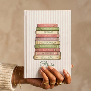 Può includere: Un diario con copertina rigida a righe verticali rosa chiaro e bianche. Al centro, una pila di nove dorsi di libri colorati con il testo: I AM LOVED, I AM BRAVE, I AM SMART, I AM KIND, I AM STRONG, I AM ENOUGH, I AM CONFIDENT, I AM BOLD, I AM BEAUTIFUL, I am Me. Il nome Olivia è stampato sotto.