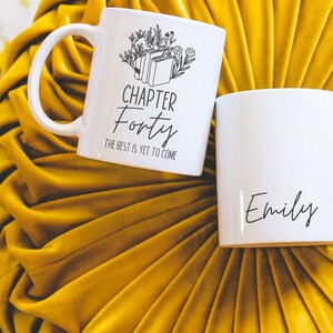 May include: Two white ceramic mugs with black text. One mug says "Chapter Forty The Best is Yet to Come" with a drawing of books and flowers. The other mug says "Emily".