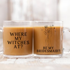 Peut inclure: Deux tasses en verre transparent avec du texte noir et des motifs. La tasse de gauche dit "Where my witches at?" La tasse de droite a un dessin au trait de mains tenant des fleurs et dit "Be my bridesmaid?"