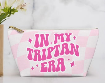 Pode incluir: Uma bolsa branca com zíper com um padrão xadrez rosa e branco. A bolsa tem um texto rosa que diz "In My Triptan Era" com estrelas rosa de cada lado do texto.
