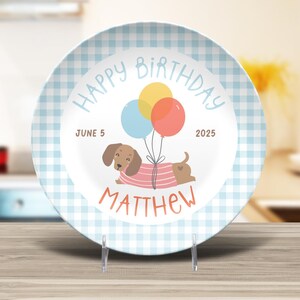 May include: A decorative plate with a light blue gingham border. The center features a dachshund wearing a pink and white striped shirt, holding balloons. Text reads "Happy Birthday," "June 5, 2025," and "Matthew."