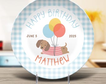 May include: A decorative plate with a light blue gingham border. The center features a dachshund wearing a pink and white striped shirt, holding balloons. Text reads "Happy Birthday," "June 5, 2025," and "Matthew."