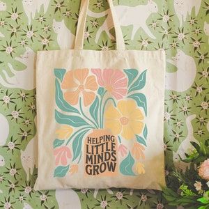 Puede incluir: Una bolsa de lona blanca con un diseño floral y el texto "Helping Little Minds Grow". La bolsa está colgada en una pared con un papel tapiz verde y blanco con un patrón repetitivo de gatos y flores.
