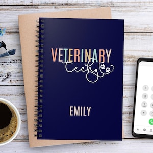 May include: A blue spiral bound notebook with the text "Veterinary Tech" in colourful letters and the name "Emily" printed below. The notebook is on a wooden surface with a cup of coffee, glasses, and a smartphone.
