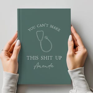 Könnte beinhalten: Ein teal-farbenes Notizbuch mit dem Text "YOU CAN'T MAKE THIS SHIT UP Amanda" und einer Stethoskop-Illustration. Das Notizbuch wird von zwei Händen vor einem hellgrauen Hintergrund gehalten.
