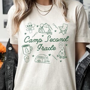 May include: A white t-shirt with a green design that says "Camp Second Grade" with camping-themed illustrations such as a tent, campfire, and a camper.