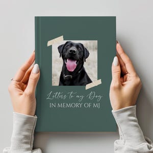 Könnte beinhalten: Ein dunkelgrünes Buch mit dem Titel "Letters to my Dog" und einem Foto eines schwarzen Labrador Retrievers. Das Foto ist mit Klebeband befestigt. Der Text "IN MEMORY OF MJ" steht unter dem Titel. Das Buch wird von einer Person gehalten.