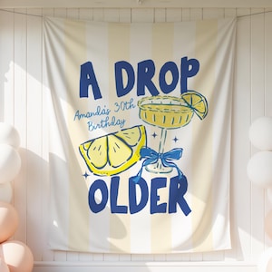 Puede incluir: Una pancarta crema con rayas amarillas, con el texto azul marino "A DROP OLDER", con una ilustración de limón y cóctel. Globos blancos y melocotón flanquean la pancarta, creando un ambiente festivo para un cumpleaños.