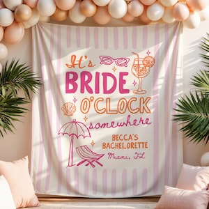 May include: A rectangular fabric backdrop featuring vertical light pink stripes and the text It's BRIDE O'CLOCK somewhere, BECCA'S BACHELORETTE, Miami, FL. Graphics include pink sunglasses, a cocktail, a seashell, a beach umbrella, and a lounge chair.