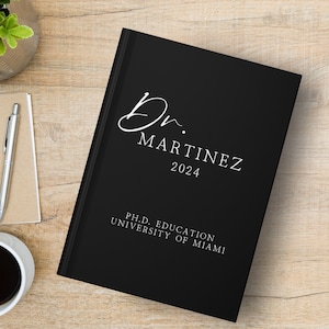 Op de afbeelding: Zwart hardback dagboek met witte tekst die "Dr. Martinez 2024" en "Ph.D. Education University of Miami" luidt.