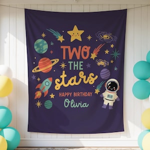 May include: A dark purple fabric backdrop featuring a space theme with planets, stars, a rocket, and an astronaut. The text reads TWO THE stars HAPPY BIRTHDAY Olivia in colorful letters. The banner is flanked by clusters of teal, yellow, and white balloons.