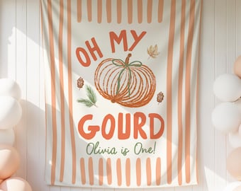 Könnte beinhalten: Ein rechteckiger Stoffhintergrund mit vertikalen orangefarbenen Streifen auf cremefarbenem Grund. In der Mitte befindet sich eine orangefarbene Kürbisillustration mit grünen Stielen, umgeben vom Text OH MY GOURD und Olivia is One! in Orange und Grün.