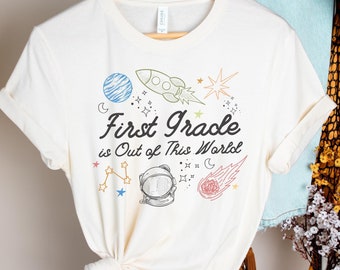 May include: A white t-shirt with a space-themed design. The shirt features a rocket, planets, stars, and an astronaut. The text on the shirt reads "First Grade is Out of This World."