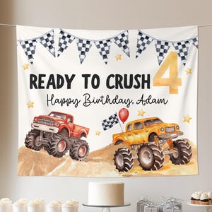 May include: A birthday party backdrop featuring two monster trucks, a red and a yellow one, on a sandy background. The banner reads "READY TO CRUSH Happy Birthday, Adam" with a large number 4. Checkered flags and star accents complete the design.