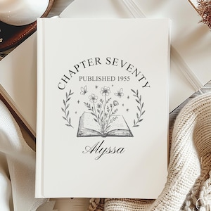 May include: A cream-coloured hardback journal with the text "Chapter Seventy Published 1955" and the name "Alyssa" in script. The cover features a floral and butterfly design. The journal is surrounded by a candle, a book, and a knitted blanket.