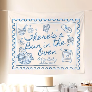 May include: A cream-colored banner with a blue border and illustrations, including a basket of bread and an oven. The banner reads "There's a Bun in the Oven, Ali's Baby Shower." A table is set with a cake, gifts, and cupcakes.