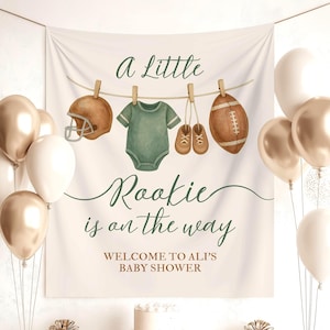 Pode incluir: Cena de ch&aacute; de beb&eacute; com uma faixa que diz "A Little Rookie is on the way". A faixa apresenta ilustra&ccedil;&otilde;es em aquarela de um capacete de futebol americano, um body, sapatos de beb&eacute; e uma bola de futebol. Bal&otilde;es dourados e brancos, presentes e um bolo.