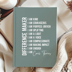 May include: A sage green journal with the words "DIFFERENCE MAKER" printed vertically on the left side. The journal's cover features a list of affirmations in white text, including "I AM KIND" and "I AM COURAGEOUS". The author's name, "Coach Thomas", is at the bottom.