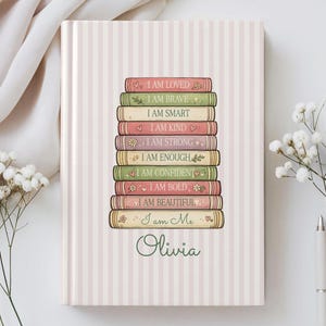 Può includere: Un diario con copertina rigida a righe verticali rosa chiaro e bianche. Al centro è raffigurata una pila di nove libri illustrati con il testo: I AM LOVED, I AM BRAVE, I AM SMART, I AM KIND, I AM STRONG, I AM ENOUGH, I AM CONFIDENT, I AM BOLD, I AM BEAUTIFUL, I am Me, e il nome Olivia.