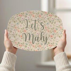 Puede incluir: Bandeja ovalada con un estampado floral y de fichas de Mahjong. La bandeja tiene un fondo beige con flores rosas y hojas verdes. Las palabras "Let's Mahj" están escritas en una fuente cursiva.