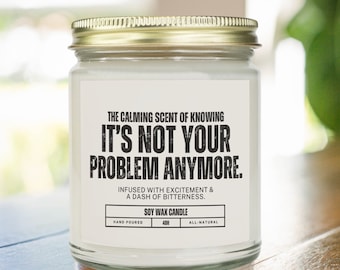 May include: A white soy wax candle with a gold lid. The label reads "The calming scent of knowing It's not your problem anymore. Infused with excitement & a dash of bitterness. Soy wax candle. Hand poured. 40h. All-natural."