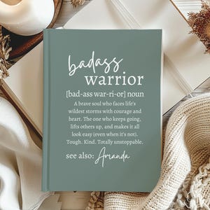 May include: A green journal with the text "badass warrior" and a definition of the word. The definition reads "[bad-ass war-ri-or] noun A brave soul who faces life's wildest storms with courage and heart. The one who keeps going, lifts others up, and makes it all look easy (even when it's not). Tough. Kind. Totally unstoppable. see also: Amanda"