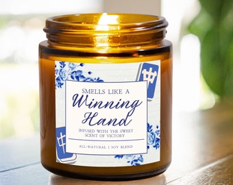 Könnte beinhalten: Ein braunes Glasgefäß mit einer Kerze und einem weißen Etikett mit der Aufschrift "Smells Like A Winning Hand" und "Infused with the sweet scent of victory." Das Etikett enthält auch ein blaues und weißes Blumenmuster und das chinesische Schriftzeichen für "Mitte".