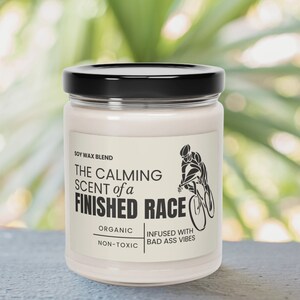 May include: A white soy wax candle with a black lid. The label reads "The Calming Scent of a Finished Race", "Soy Wax Blend", "Organic", "Non-Toxic", "Infused with Bad Ass Vibes". A black and white illustration of a cyclist is on the label.