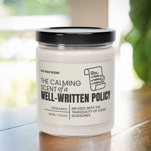 May include: A white candle with a black lid and a label that reads "The Calming Scent of a Well-Written Policy". The label also says "Soy Wax Blend", "Organic", "Non-Toxic", "Infused with the Tranquility of Clear Guidelines".