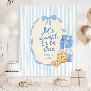 Może przedstawiać: Niebiesko-biały sztandar w paski z napisem "It's Sweet to be One" i "Adam's 1st Birthday". Na sztandarze znajduje się karton mleka, ciasteczka i niebieski wzór wstążki. Białe i złote balony w tle.