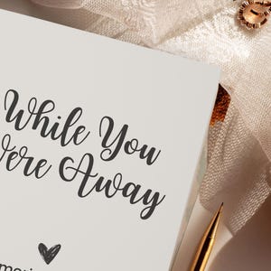 May include: A white journal with the text "While You Were Away" in black cursive script. A small heart is drawn below the text. The journal is on a white surface with a gold pen and a cup of tea.
