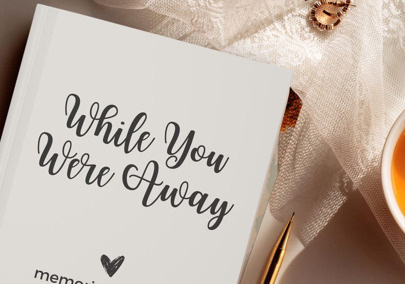 May include: A white journal with the text "While You Were Away" in black cursive script. A small heart is drawn below the text. The journal is on a white surface with a gold pen and a cup of tea.